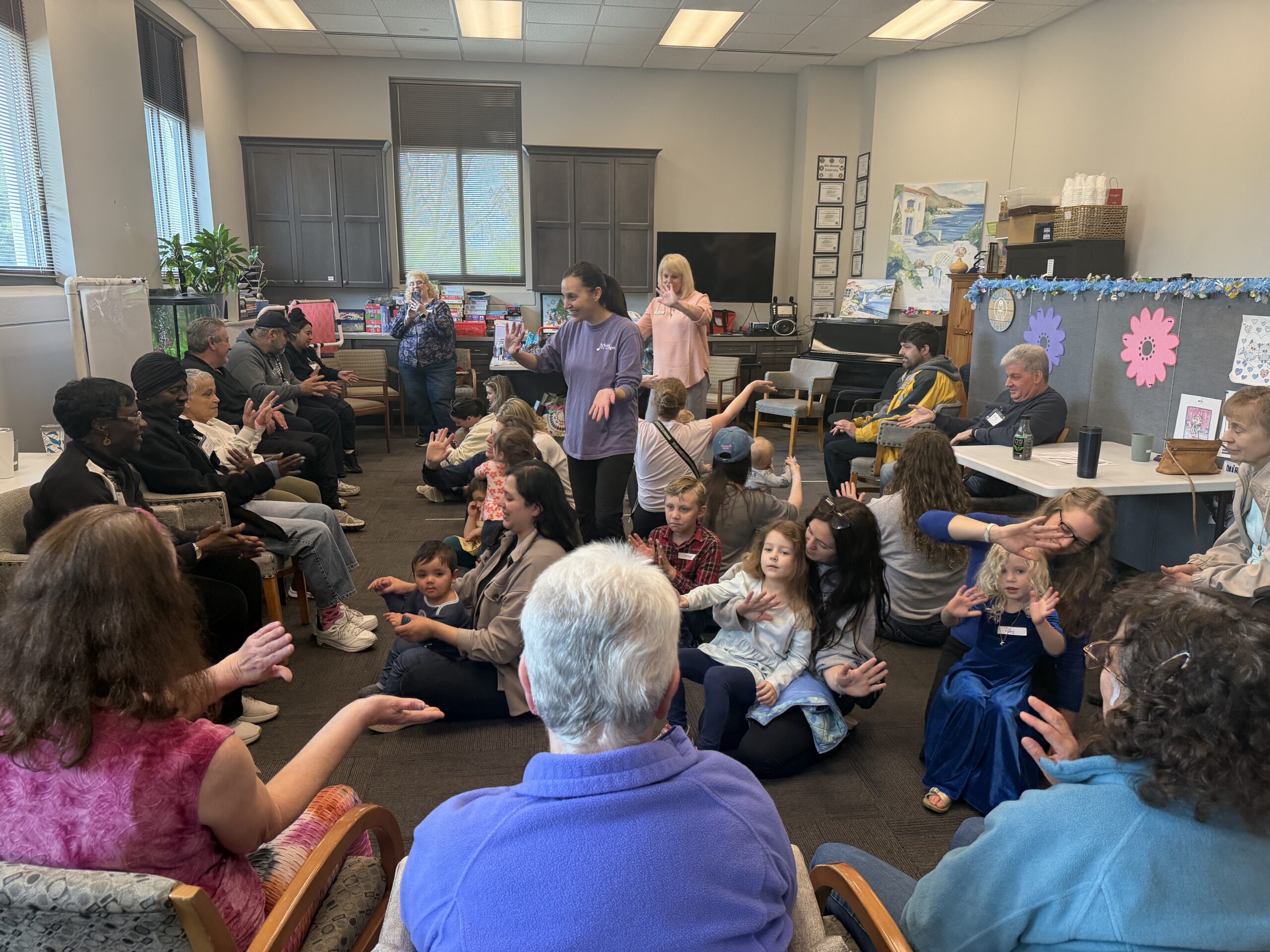 A group of children and adults sit in a circle in a classroom, with many people using sign language to communicate.