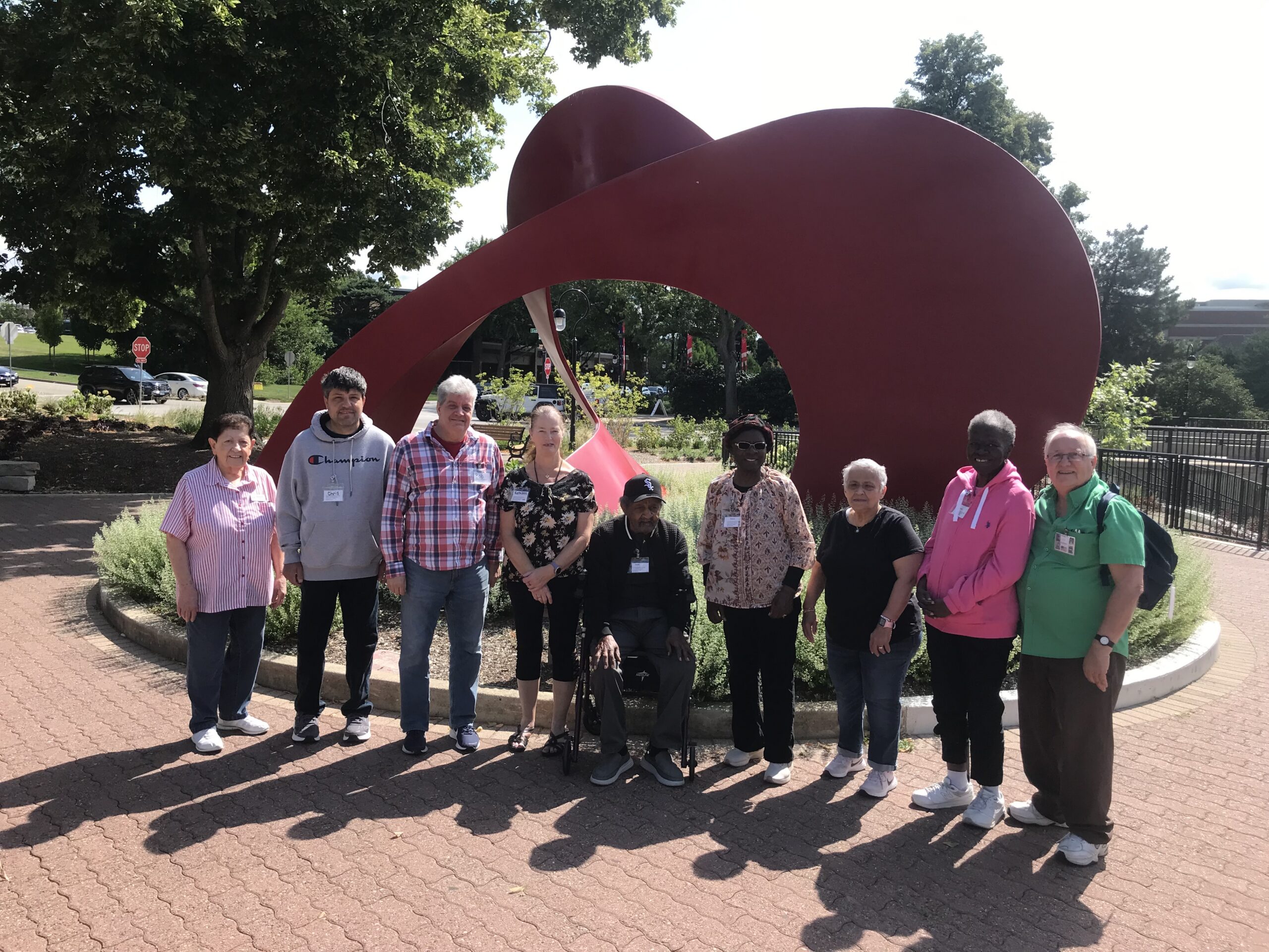 A group of nine people standing together outdoors in front of a large, curved red metal sculpture.