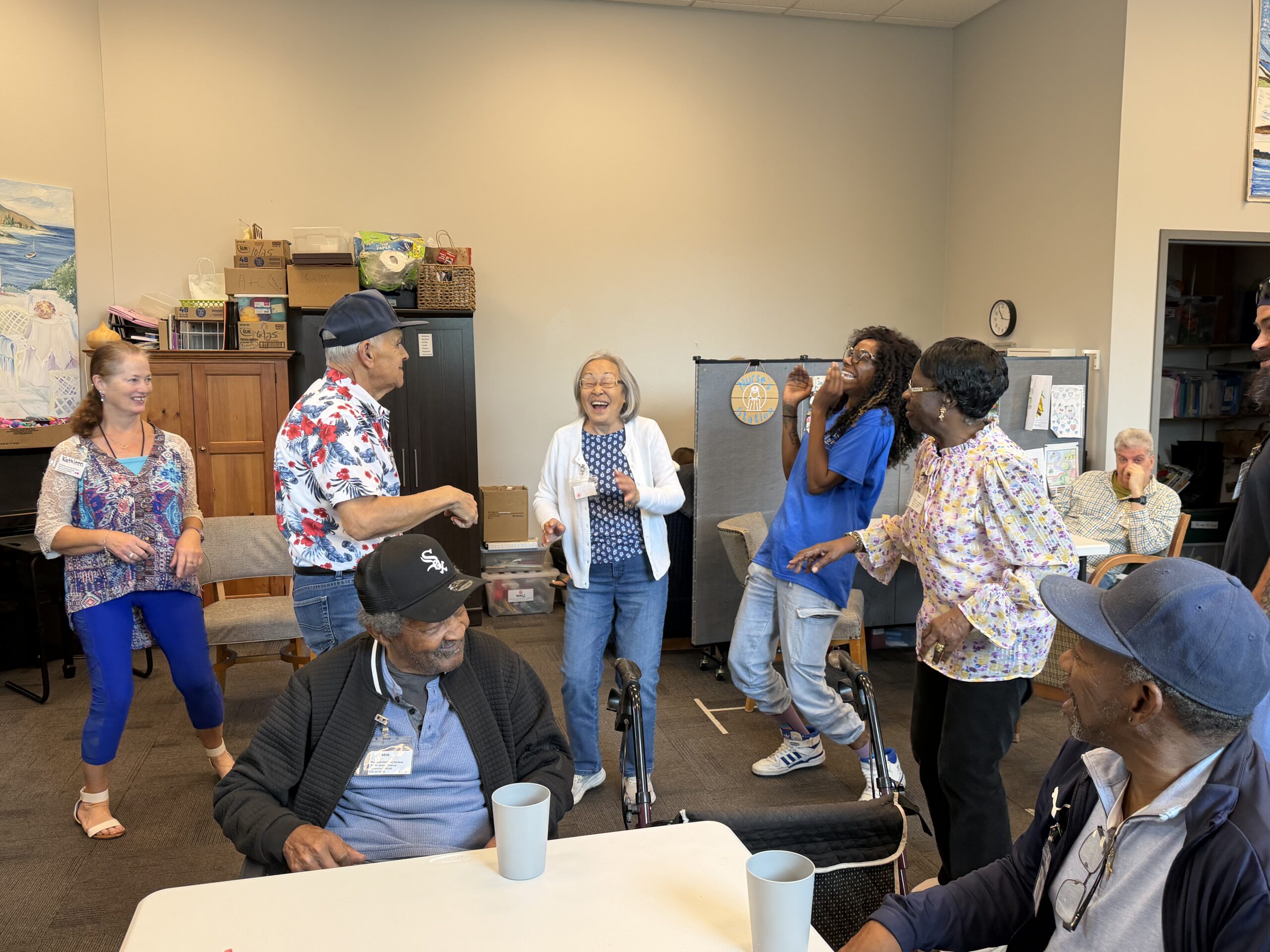 A group of diverse people in a room laughing and interacting, with two men seated at a table in the foreground.
