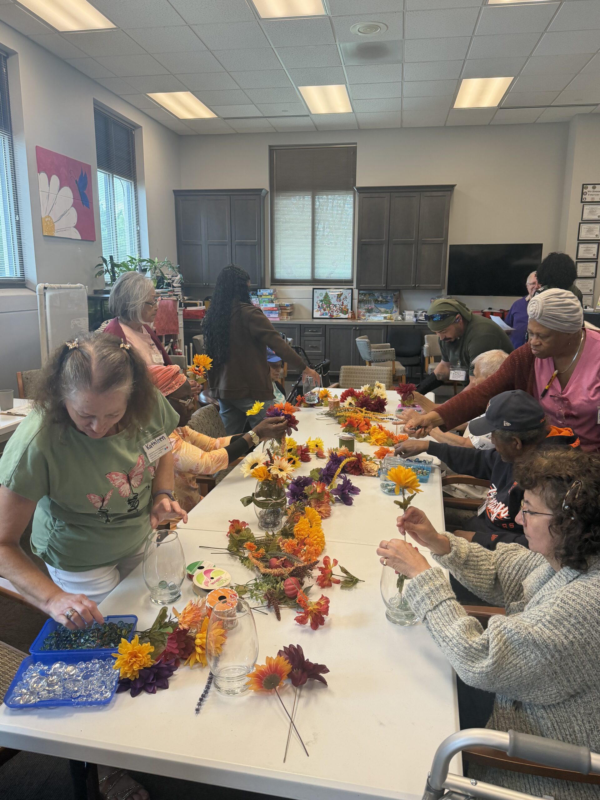 A group of people sitting around a long table creating floral arrangements with silk flowers and glass vases.
