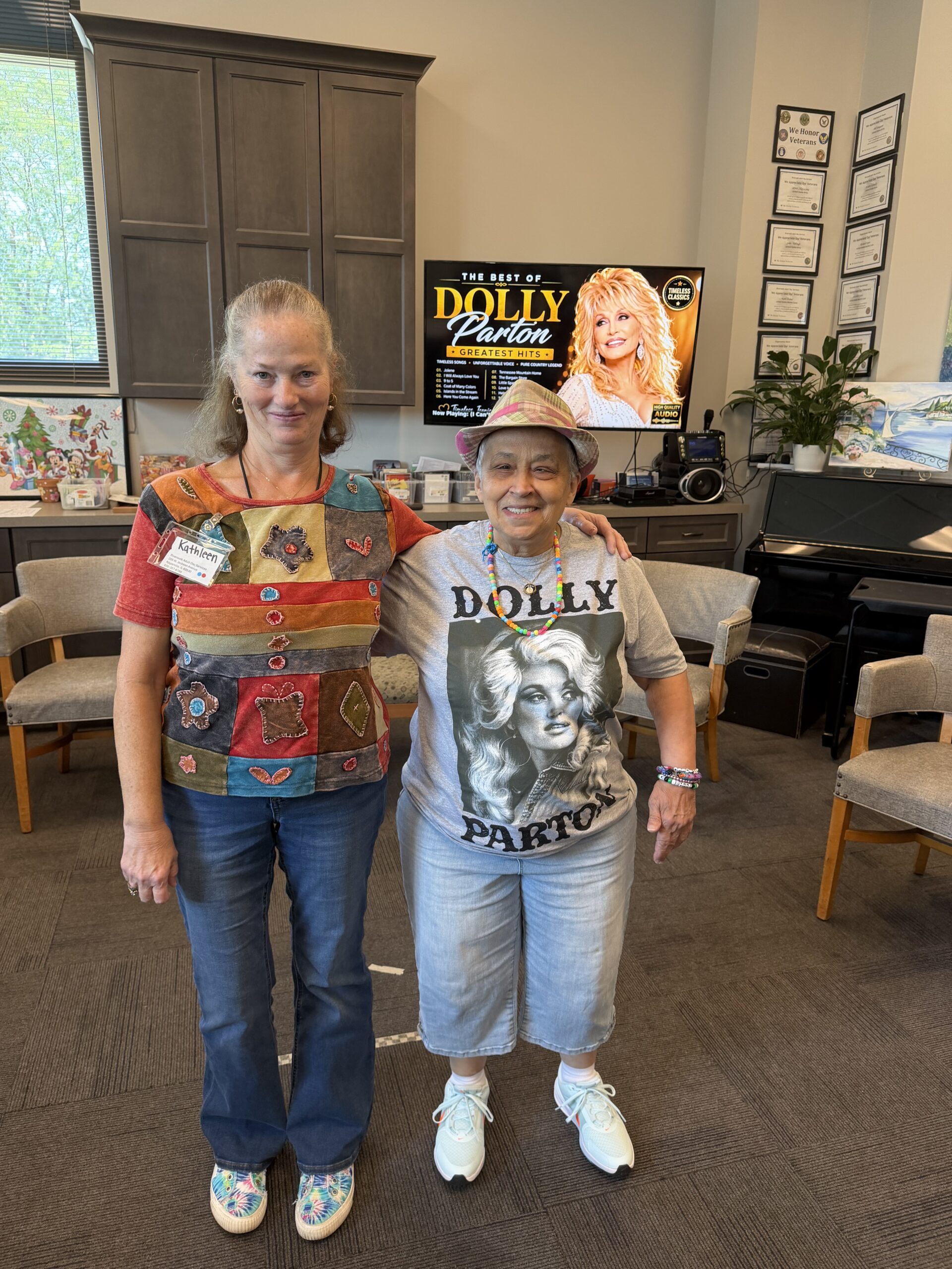 Two women stand arm-in-arm in front of a television displaying a Dolly Parton concert, one wearing a colorful patchwork shirt and the other a grey Dolly Parton graphic t-shirt and fedora.