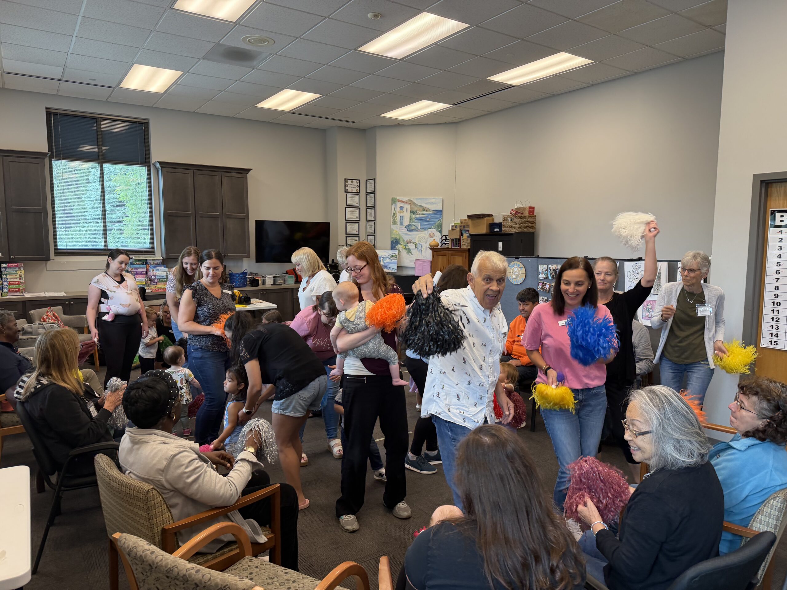 A group of people of various ages, including a baby, hold colorful pom-poms while gathered in a community room.